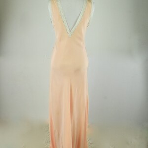 May include: A peach-colored, floor-length slip dress on a mannequin. The dress features a V-neckline trimmed with white lace and a flowing, silky fabric. The dress is sleeveless and has a simple, elegant design.