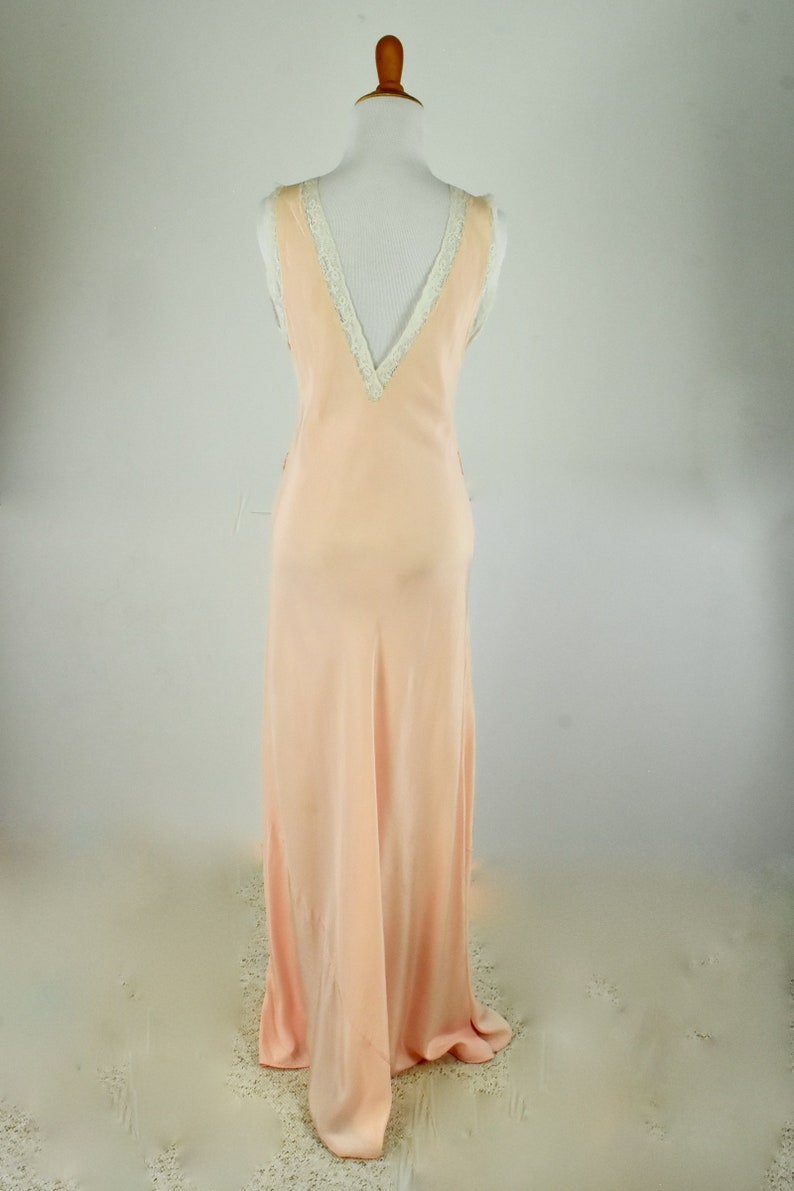 May include: A peach-colored, floor-length slip dress on a mannequin. The dress features a V-neckline trimmed with white lace and a flowing, silky fabric. The dress is sleeveless and has a simple, elegant design.