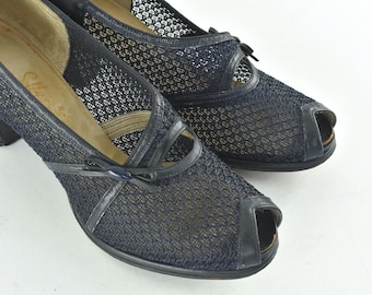 May include: A pair of vintage navy blue peep-toe heels. The shoes feature a mesh design with leather accents and small bows. The heels are a dark navy color, and the soles are a light tan color. The shoes are likely from the mid-20th century.