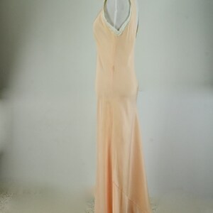 May include: A peach-colored, floor-length gown with a V-neckline and a flowing skirt. The dress has a simple, elegant design with a subtle sheen. The neckline is trimmed with delicate white lace. The dress is displayed on a mannequin.