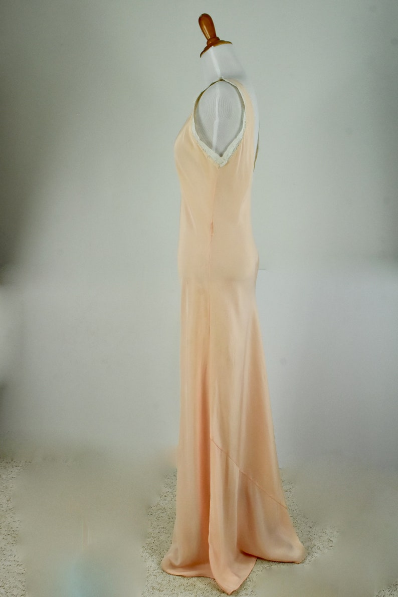 May include: A peach-colored, floor-length gown with a V-neckline and a flowing skirt. The dress has a simple, elegant design with a subtle sheen. The neckline is trimmed with delicate white lace. The dress is displayed on a mannequin.