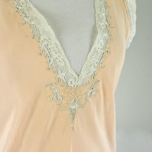 May include: A peach-colored, vintage-style top with a V-neckline. The neckline and short sleeves are trimmed with delicate, off-white lace. The center of the neckline features an embroidered design with small, decorative beads.