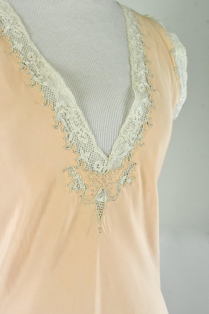 May include: A peach-colored, vintage-style top with a V-neckline. The neckline and short sleeves are trimmed with delicate, off-white lace. The center of the neckline features an embroidered design with small, decorative beads.