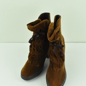 Puede incluir: Un par de botas de terciopelo marrón con tacón alto. Las botas tienen un diseño arrugado en la parte superior y están atadas con una cinta marrón. Las botas están sobre una superficie blanca.