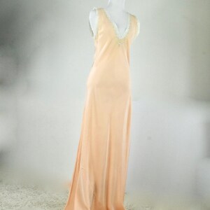 May include: A peach-colored, floor-length slip dress on a mannequin. The dress features a V-neckline trimmed with white lace and delicate embellishments. The fabric drapes smoothly, creating a flowing silhouette.