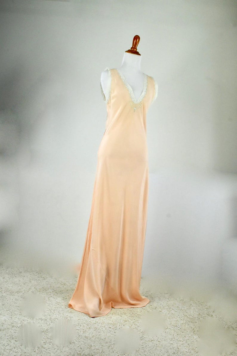 May include: A peach-colored, floor-length slip dress on a mannequin. The dress features a V-neckline trimmed with white lace and delicate embellishments. The fabric drapes smoothly, creating a flowing silhouette.