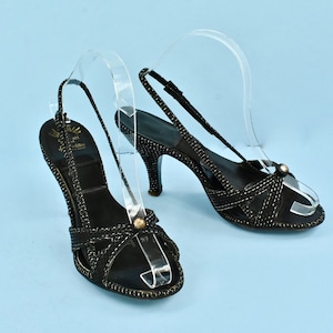 May include: Black slingback heels with a stiletto heel. The shoes feature a crisscross design with white stitching and a small gold-colored accent on the toe. The heels are displayed on a light blue background.