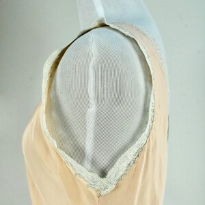 May include: A peach-colored slip dress with a delicate white lace trim along the neckline and armholes. The dress is displayed on a white mannequin, showcasing its simple, elegant design. The fabric appears soft and lightweight.