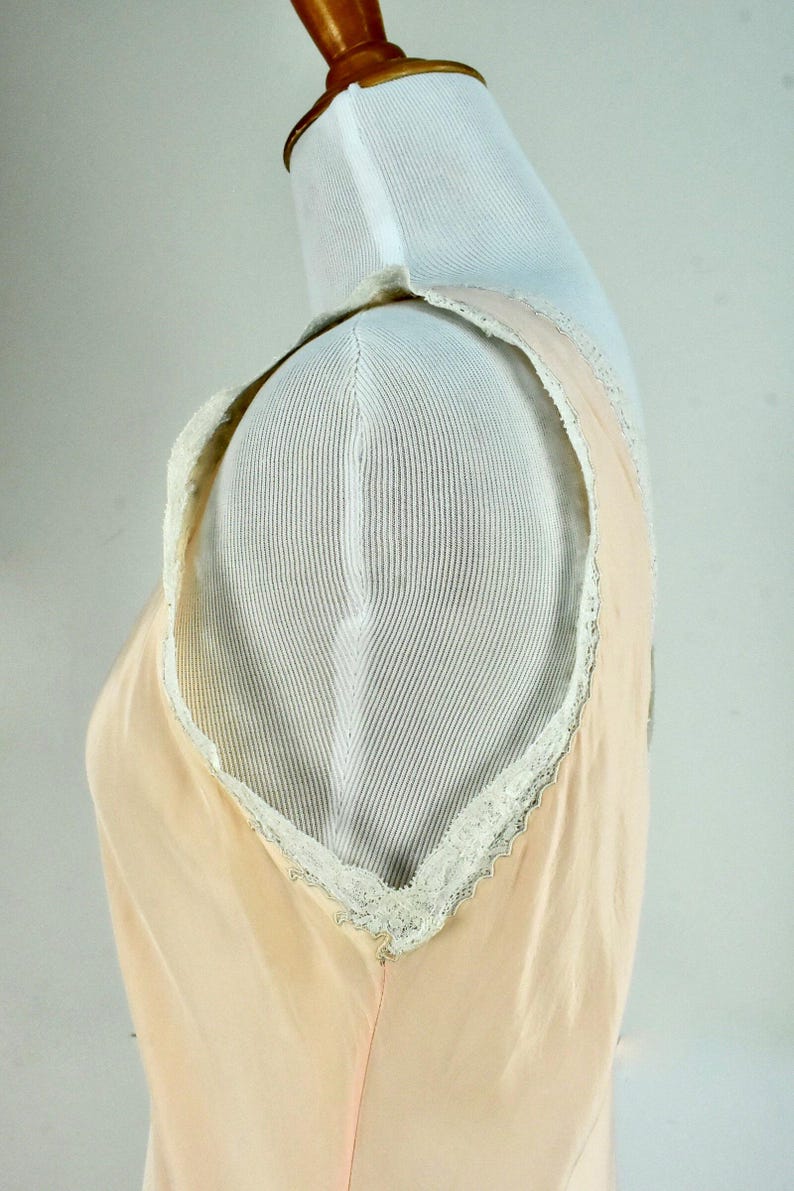 May include: A peach-colored slip dress with a delicate white lace trim along the neckline and armholes. The dress is displayed on a white mannequin, showcasing its simple, elegant design. The fabric appears soft and lightweight.