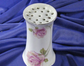May include: White ceramic flower frog with a decorative floral design featuring pink roses. The frog has a circular top with multiple holes for holding flower stems.