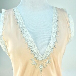 May include: A peach-colored vintage slip with a V-neckline, trimmed with delicate white lace. The lace detailing extends along the neckline and around the shoulders, creating an elegant and feminine look. The slip is displayed on a mannequin.