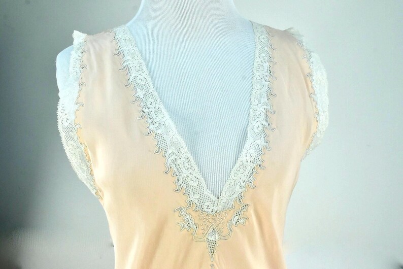 May include: A peach-colored vintage slip with a V-neckline, trimmed with delicate white lace. The lace detailing extends along the neckline and around the shoulders, creating an elegant and feminine look. The slip is displayed on a mannequin.