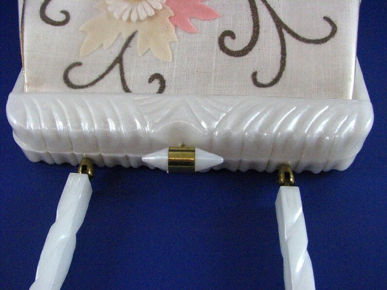 May include: A white vintage clutch purse with a floral design and gold-tone hardware. The purse has a white, textured exterior and two white handles.