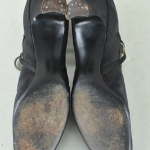 May include: A pair of black satin heeled shoes with a buckle strap detail. The shoes have a pointed toe and a high heel. The soles of the shoes are worn and show signs of use.