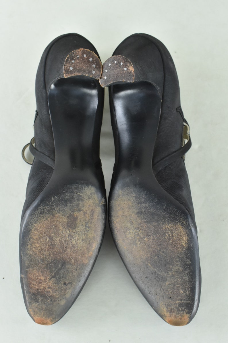May include: A pair of black satin heeled shoes with a buckle strap detail. The shoes have a pointed toe and a high heel. The soles of the shoes are worn and show signs of use.