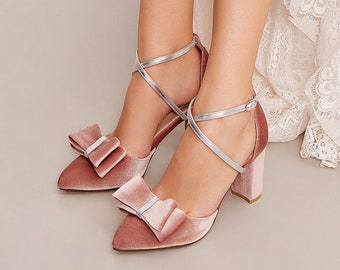 May include: A pair of blush pink velvet heels with silver straps and large velvet bows. The heels have a block heel and a pointed toe.