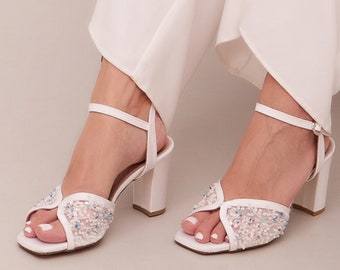 May include: White heeled sandals with a delicate beaded design. The sandals have a square toe and a block heel.