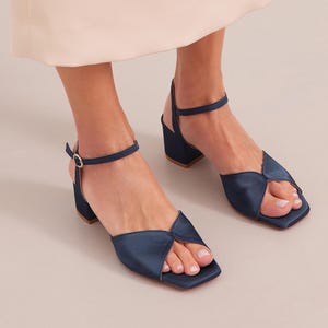 May include: Navy blue satin heeled sandals with a square toe and an ankle strap. The sandals have a low block heel.
