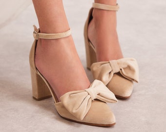 Può includere: Scarpe con tacco in camoscio beige con un grande fiocco sulla punta. Le scarpe hanno un cinturino alla caviglia con chiusura a fibbia.
