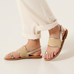 May include: Gold leather sandals with a woven strap and a buckle closure. The sandals have a flat sole and a toe ring.