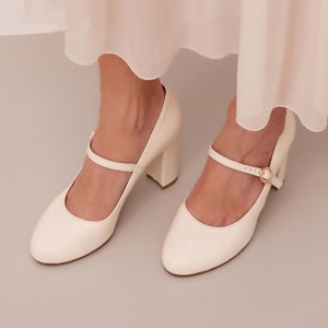 May include: A pair of off-white leather heeled shoes with a buckle strap. The shoes have a rounded toe and a block heel.