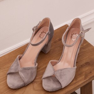 May include: A pair of gray suede heeled sandals with ankle straps and a buckle closure. The sandals have a low block heel and an open toe design. The brand name "Greek Chic" is visible on the insole.