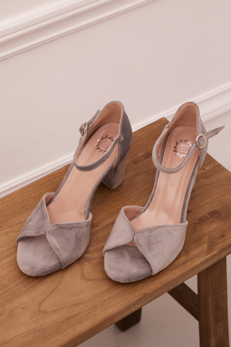 May include: A pair of gray suede heeled sandals with ankle straps and a buckle closure. The sandals have a low block heel and an open toe design. The brand name "Greek Chic" is visible on the insole.