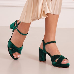 May include: A pair of green velvet heeled sandals with ankle straps. The sandals have a criss-cross design on the front and a block heel.
