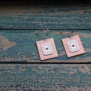 May include: A pair of pink and silver earrings with clock face designs. The clock faces have Roman numerals and a red dot in the centre.