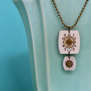 Puede incluir: Un collar de cadena dorada con un colgante blanco que presenta tres esferas de reloj con números romanos y engranajes. El collar se muestra sobre un fondo azul claro.