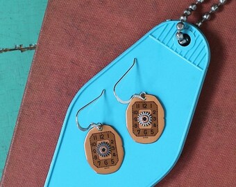 Peut inclure: Une paire de boucles d'oreilles sur un fond bleu. Les boucles d'oreilles sont en métal et ont un design de cadran d'horloge avec des chiffres romains et un engrenage au centre. Les boucles d'oreilles sont sur une étiquette de clé de motel bleue.