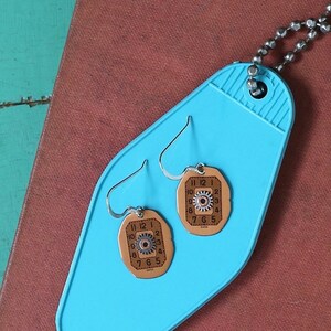 Puede incluir: Un par de pendientes con un fondo azul. Los pendientes están hechos de metal y tienen un diseño de esfera de reloj con números romanos y un engranaje en el centro. Los pendientes están en una etiqueta de llave de motel azul.