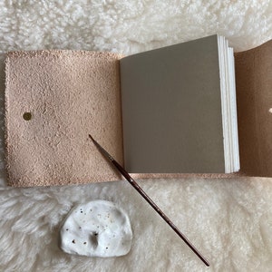 May include: A light pink leather journal, open to reveal a stack of blank pages. A small, white ceramic paint palette and a thin paintbrush are placed beside the journal. The journal has a brass snap closure.