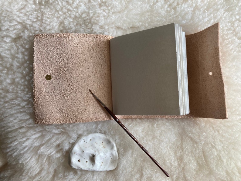 May include: A light pink leather journal, open to reveal a stack of blank pages. A small, white ceramic paint palette and a thin paintbrush are placed beside the journal. The journal has a brass snap closure.