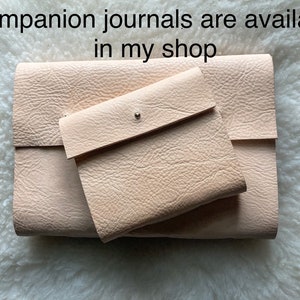 May include: A set of light beige leather journals and a small matching pouch. The journals are rectangular with a textured surface. The pouch has a flap closure with a silver snap. Text at the top says "Companion journals are available in my shop."