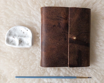 May include: A brown leather journal with a brass button closure, a small white ceramic dish, and a blue paintbrush with a gold ferrule. The items are arranged on a white, fluffy surface.