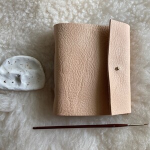 May include: A light beige leather journal with a press stud closure, a small white ceramic dish, and a thin paintbrush with a dark brown handle. The items are arranged on a textured, off-white surface.