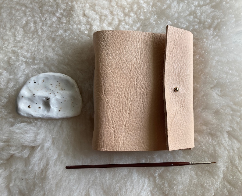 May include: A light beige leather journal with a press stud closure, a small white ceramic dish, and a thin paintbrush with a dark brown handle. The items are arranged on a textured, off-white surface.