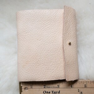May include: A small, light beige leather journal with a textured surface. The journal is closed with a flap and a gold-coloured snap. A wooden ruler with the text "One Yard" is placed next to the journal.