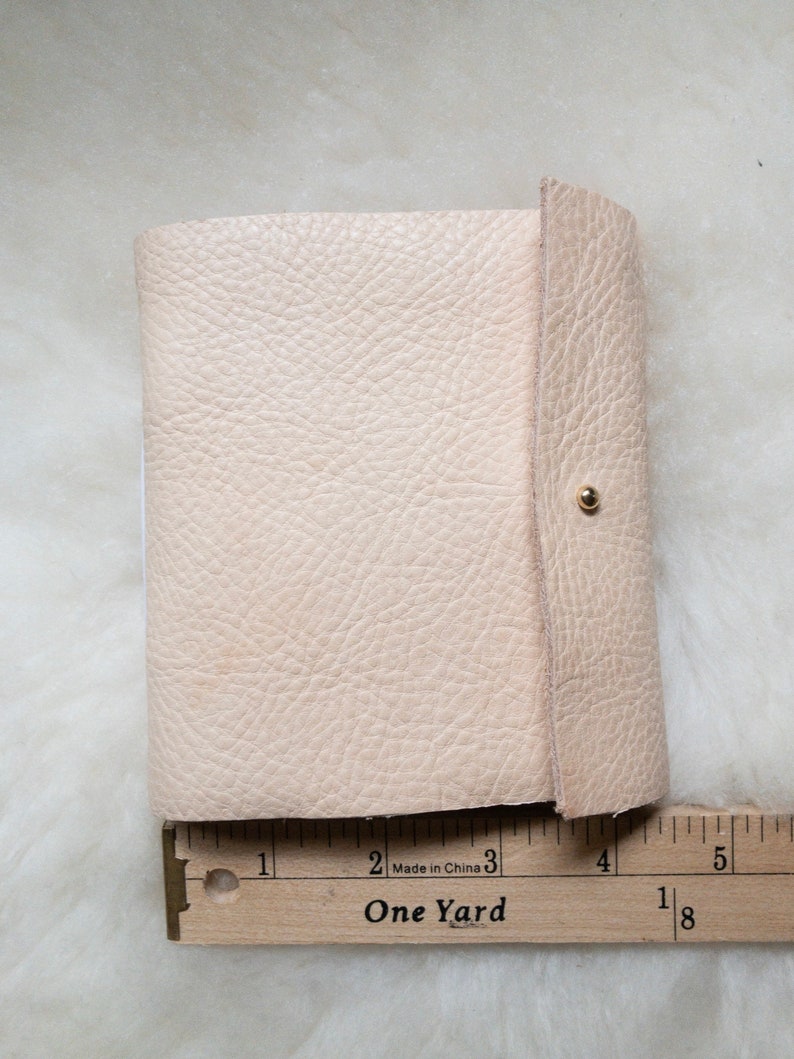 May include: A small, light beige leather journal with a textured surface. The journal is closed with a flap and a gold-coloured snap. A wooden ruler with the text "One Yard" is placed next to the journal.