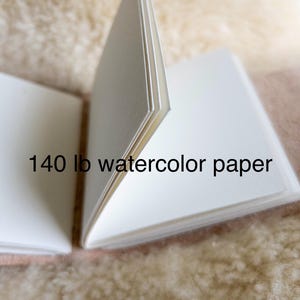 May include: An open sketchbook with bright white watercolour paper, displaying the text "140 lb watercolour paper". The pages are fanned open, showing the paper's thickness. The background is a soft, textured, beige surface.