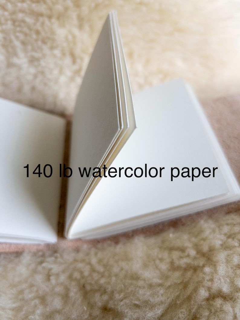 May include: An open sketchbook with bright white watercolour paper, displaying the text "140 lb watercolour paper". The pages are fanned open, showing the paper's thickness. The background is a soft, textured, beige surface.