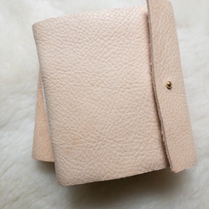 May include: A light beige leather journal with a textured surface. The journal is closed with a flap and a gold-coloured snap. The pages are visible on the left side of the journal.