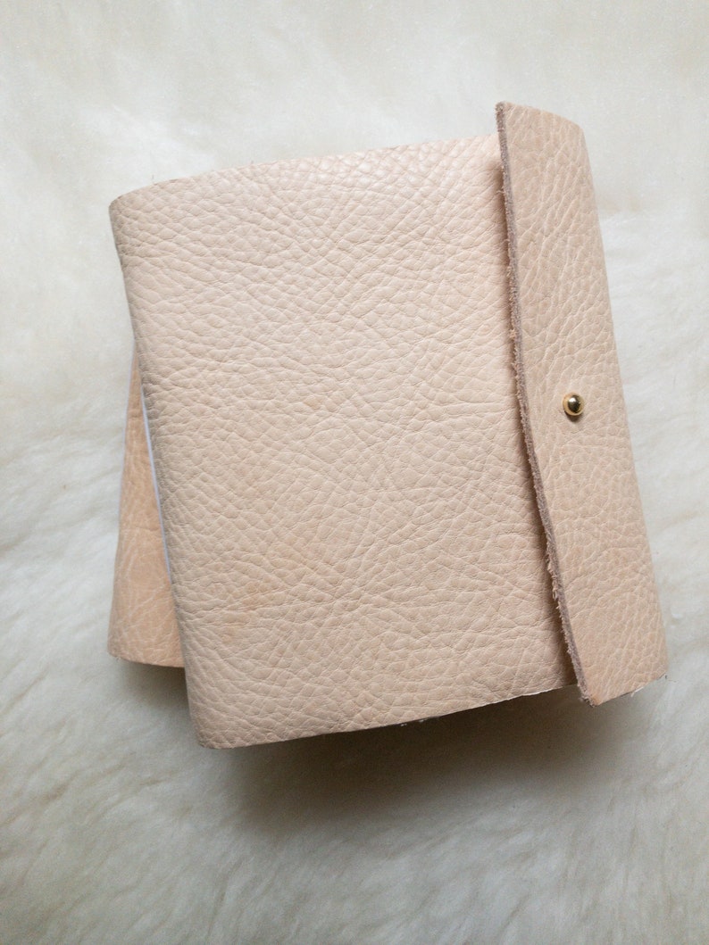 May include: A light beige leather journal with a textured surface. The journal is closed with a flap and a gold-coloured snap. The pages are visible on the left side of the journal.