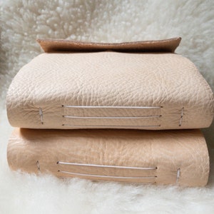 May include: Two stacked, light beige leather journals with visible stitching. The top journal has a folded-over flap. The journals are resting on a fluffy, white surface, creating a soft, neutral aesthetic.