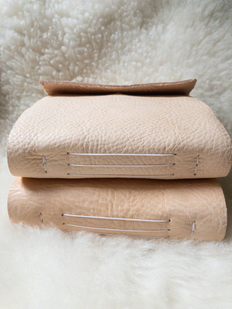 May include: Two stacked, light beige leather journals with visible stitching. The top journal has a folded-over flap. The journals are resting on a fluffy, white surface, creating a soft, neutral aesthetic.