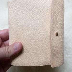 May include: A light beige leather journal with a textured surface. The journal is held open, revealing the interior pages. A gold-coloured snap closure secures the cover.