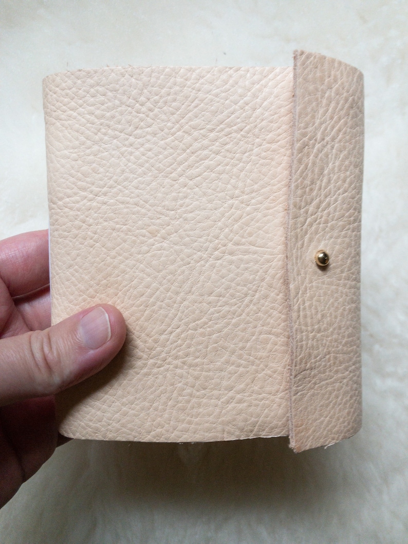 May include: A light beige leather journal with a textured surface. The journal is held open, revealing the interior pages. A gold-coloured snap closure secures the cover.