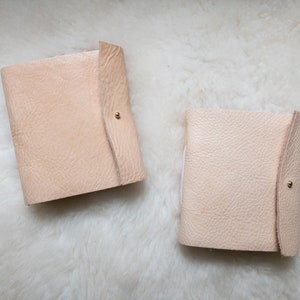 May include: Two light beige leather journals with a textured surface and a flap closure secured by a small gold-coloured stud. The journals are rectangular and appear to be handmade. The background is a white, fluffy surface.