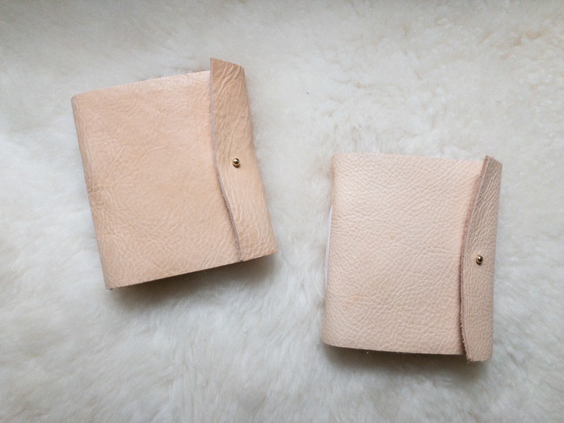 May include: Two light beige leather journals with a textured surface and a flap closure secured by a small gold-coloured stud. The journals are rectangular and appear to be handmade. The background is a white, fluffy surface.